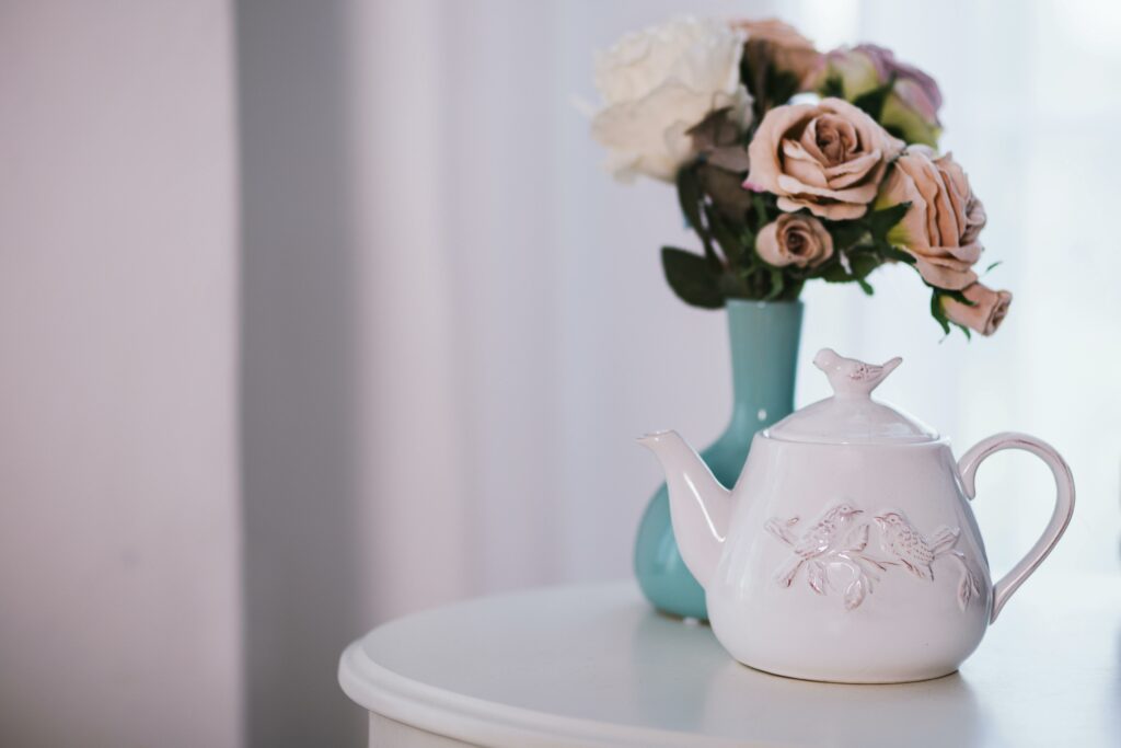 A beautiful white teapot with bird motifs beside a pastel bouquet in a blue vase indoors.